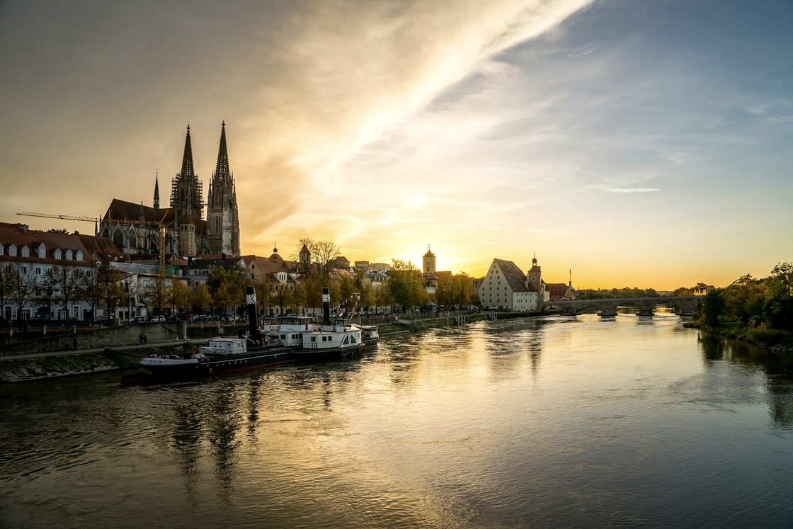 Regensburg an der Donau im Abendlicht: die Türme des Doms über der Altstadt, ein Schiff am Ufer, die Sonne tief über dem Fluss.