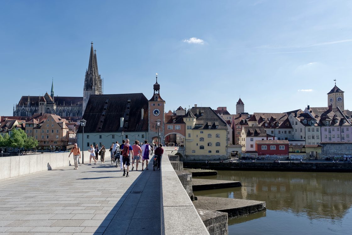 Fußgänger und Radfahrer überqueren die Steinerne Brücke Richtung Altstadt, dahinter der Brücktorturm und die Türme des Regensburger Doms.