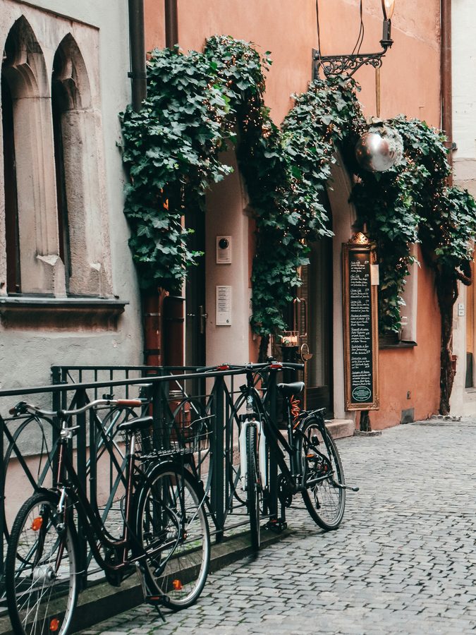 Fahrräder an einer efeubewachsenen Hauswand in einer Regensburger Altstadtgasse, daneben ein gotisches Fenster und Kopfsteinpflaster.